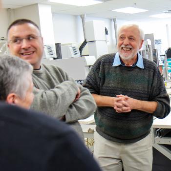 A professor stading in a biology lab surrounded by machinery, while others standby in preparation for a talk