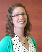 Holly Bates wearing glasses, a green sweater and white shirt with spots in front of a reddish brown background, smiling at the camera.