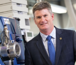Neil Emery standing in a lab next to a piece of equipment, wearing a sports jacket, checkered shirt and a royal blue tie, smiling at the camera.