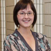 Sarah West standing in front of tiled walls wearing glasses and a mottled teal, cream, and grey shirt, smiling at the camera.