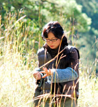 Susan Chow standing in a field of tall grasses with trees behind her on a sunny day, looking down at her hands, identifying a plant.