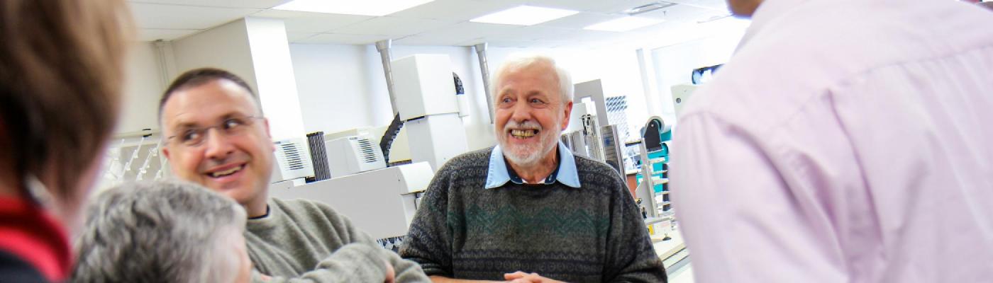A professor stading in a biology lab surrounded by machinery, while others standby in preparation for a talk
