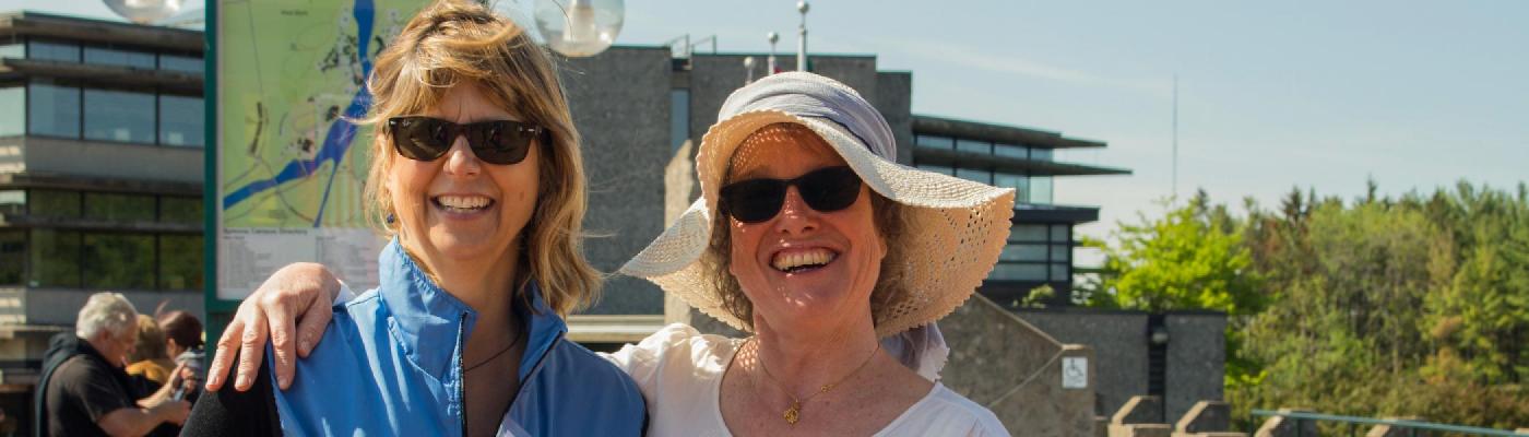 2 women in sunglasses, smiling at the camera, stadning side by side outsie of the Bata library on a sunny afternoon