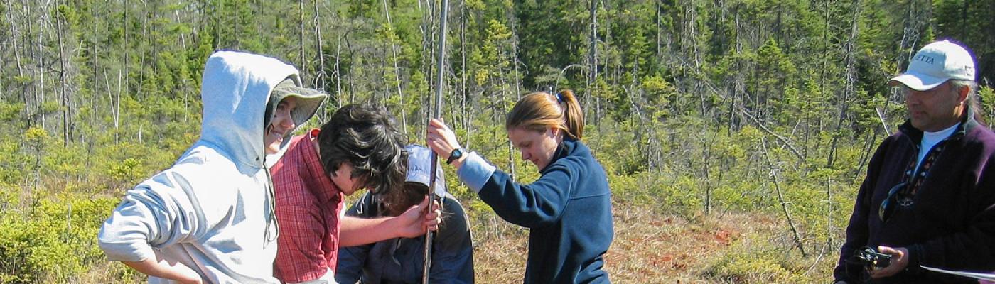 alt="A group of students standing in a wetland taking a sample of earth, dressed in summer clothes"