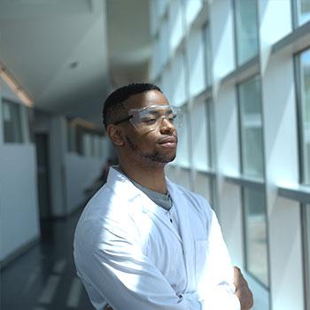 Photo of man in lab coat and safety goggles looking through a window